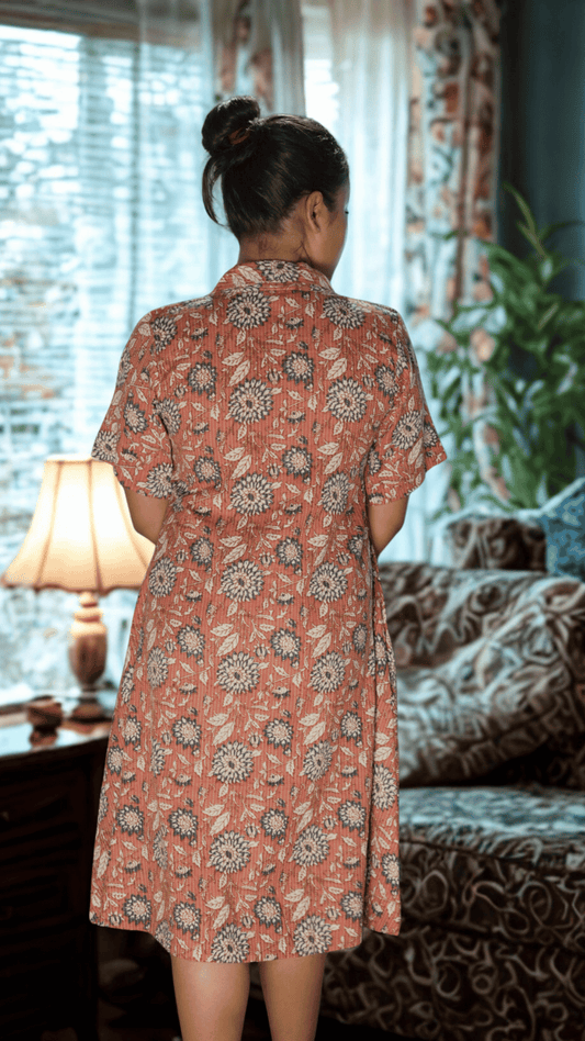 Woman wearing a patterned dress standing in a room with curtains and a lamp.