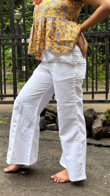 Person wearing a yellow floral top and white pants standing outdoors with a metal fence and greenery in the background.
