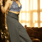 Woman in a blue patterned top and gray wide-leg pants standing in a room with warm lighting.