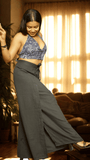Woman in a blue patterned top and gray wide-leg pants standing in a room with warm lighting.