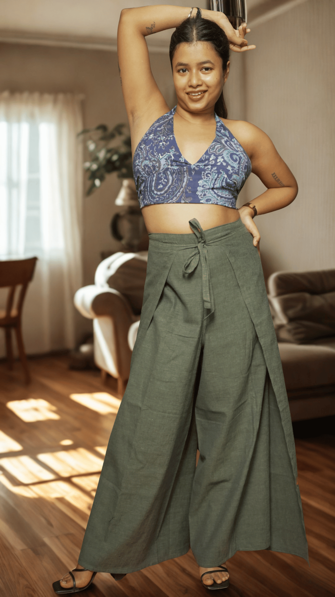Woman wearing a blue patterned top and green wide-leg pants in a living room.