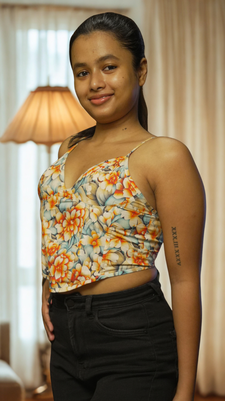 Woman wearing a floral top and black pants in a room with curtains and a lamp.