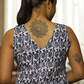 Woman wearing a blue patterned dress with tattoos visible on her back and arm, standing in a room with curtains.